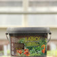 Ein 1 kg-Plastikbehälter von La'Bio | Kräuter- & Gemüsedünger, auf dessen Etikett Tomaten und Paprika abgebildet sind, steht auf einer Holzfläche mit unscharfem Hintergrund.