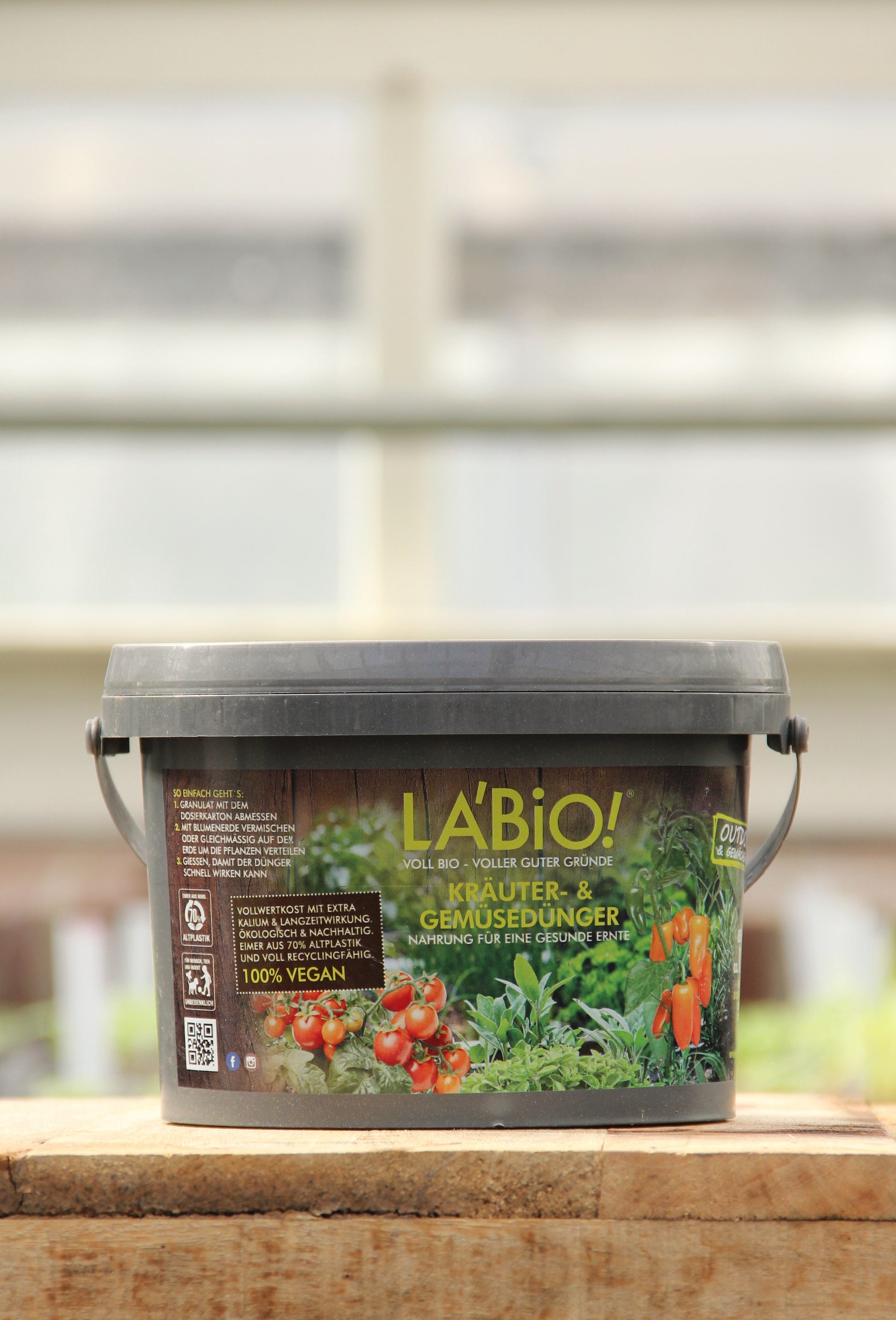 Ein 1 kg-Plastikbehälter von La'Bio | Kräuter- & Gemüsedünger, auf dessen Etikett Tomaten und Paprika abgebildet sind, steht auf einer Holzfläche mit unscharfem Hintergrund.