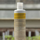 Eine aufrecht stehende weiße Kunststoffflasche von AMN 'Fusek' für kräftige Pflanzenzellen 0,5 l mit deutscher Aufschrift und der Angabe von biologischem Pflanzenhilfsmittel und Mikroorganismen steht auf Holz mit unscharfen grünen Pflanzen und einem Gewächshaus im Hintergrund.