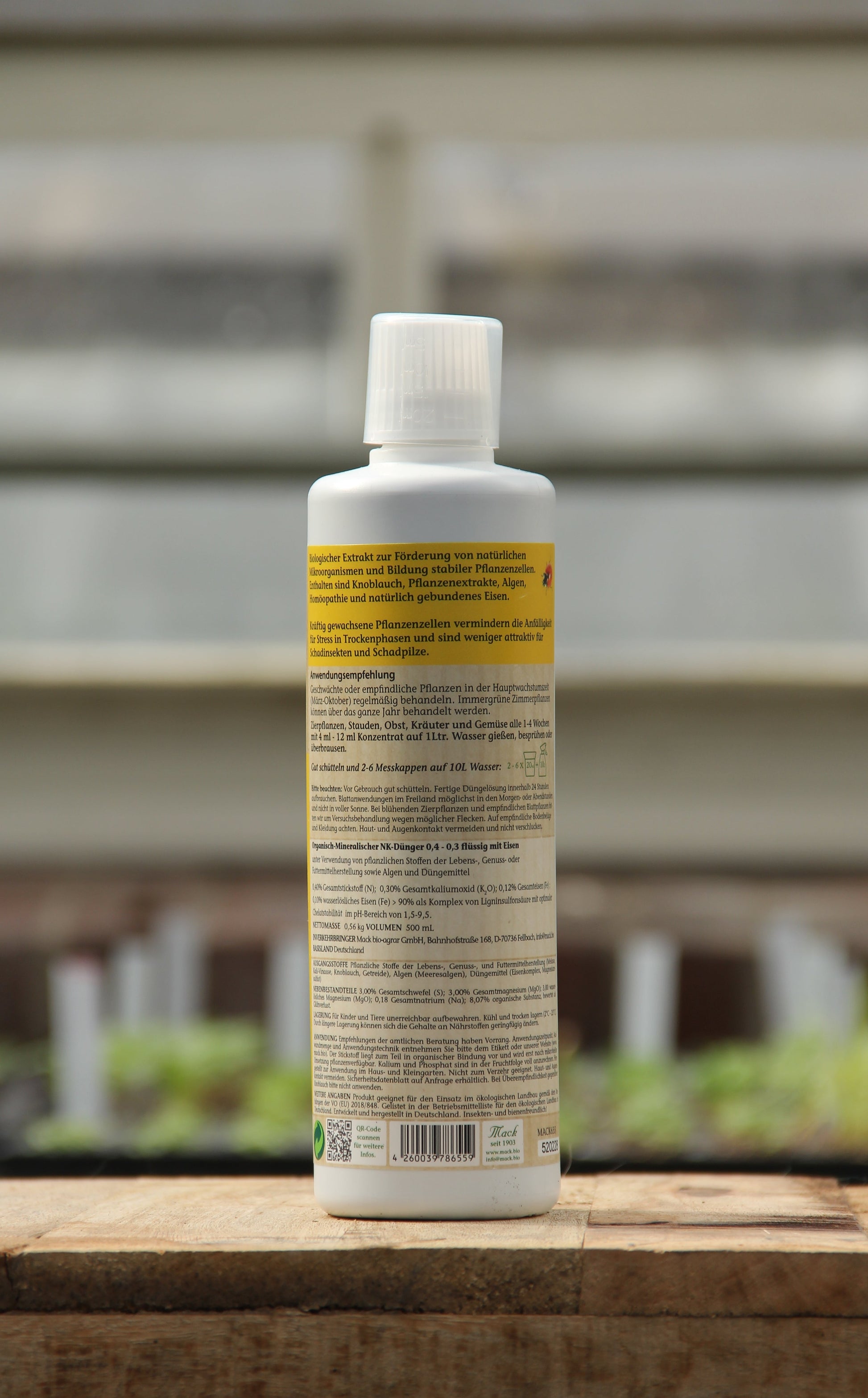 Eine aufrecht stehende weiße Kunststoffflasche von AMN 'Fusek' für kräftige Pflanzenzellen 0,5 l mit deutscher Aufschrift und der Angabe von biologischem Pflanzenhilfsmittel und Mikroorganismen steht auf Holz mit unscharfen grünen Pflanzen und einem Gewächshaus im Hintergrund.