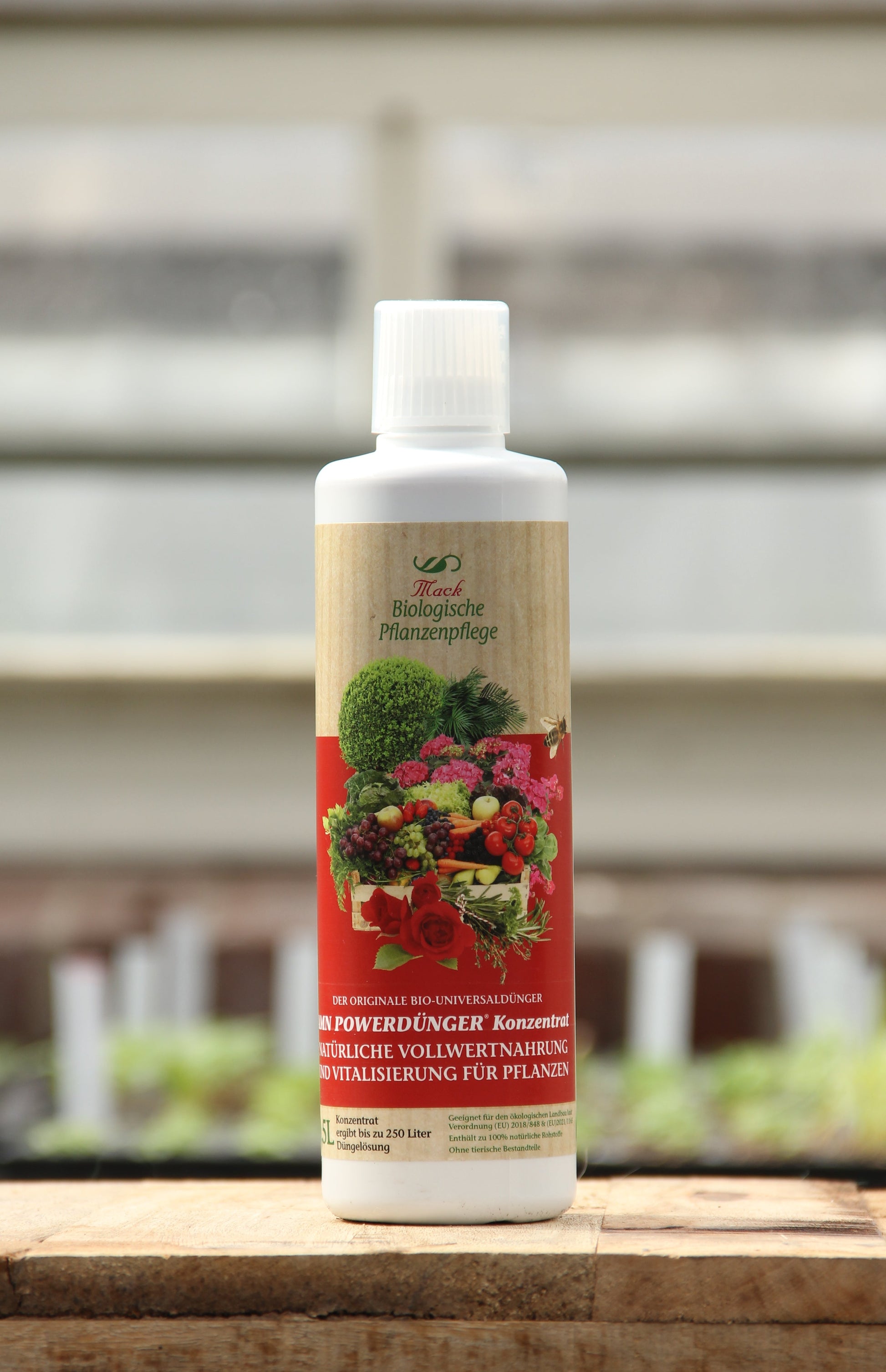 Eine weiße 0,5-Liter-Flasche des organischen Düngers "Powerdünger" von AMN mit einem roten Etikett, auf dem Obst und Gemüse abgebildet sind, steht auf einer Holzfläche, der Garten ist unscharf im Hintergrund zu sehen.