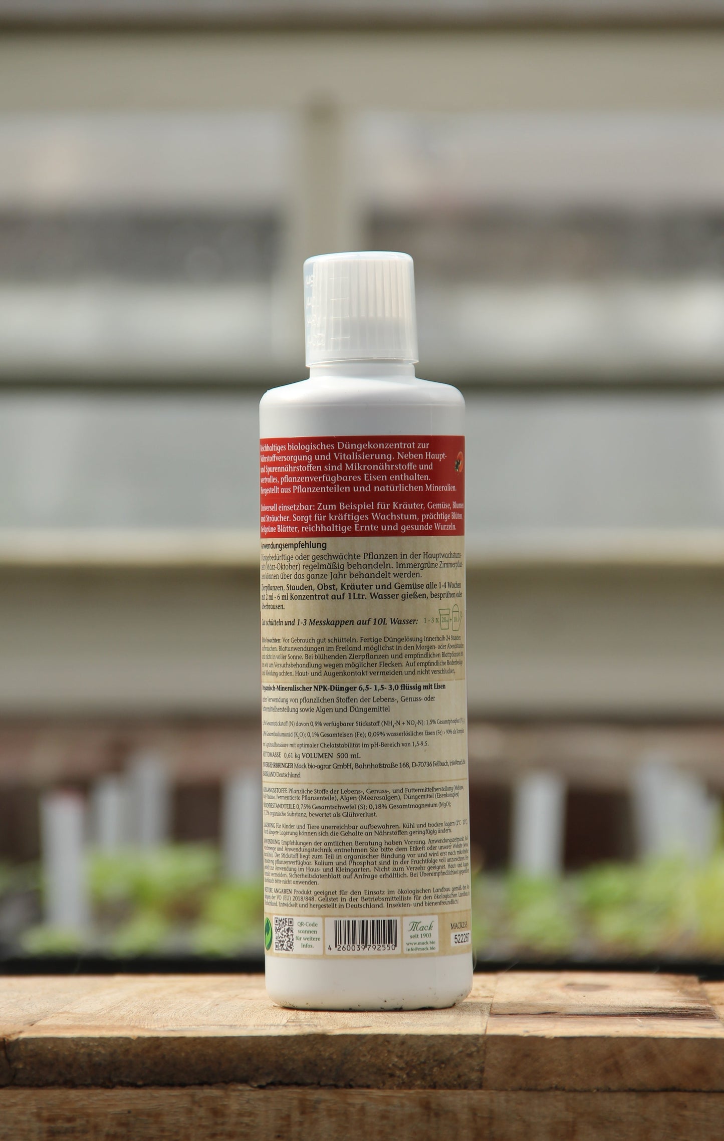Eine weiße Plastikflasche von AMN |'Powerdünger' 0,5 l mit einem roten und beigen deutschen Etikett steht aufrecht auf Holz, mit verschwommenen grünen Pflanzen und einer Gewächshausstruktur im Hintergrund.