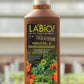 Eine braune 1-Liter-Plastikflasche von LaBio | Kräuter & Gemüsedünger mit einem grünen Etikett, auf dem Tomaten und Kräuter abgebildet sind, im Freien mit unscharfen Pflanzen im Hintergrund.