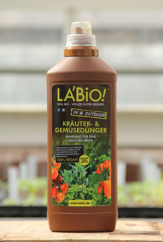 Eine braune 1-Liter-Plastikflasche von LaBio | Kräuter & Gemüsedünger mit einem grünen Etikett, auf dem Tomaten und Kräuter abgebildet sind, im Freien mit unscharfen Pflanzen im Hintergrund.
