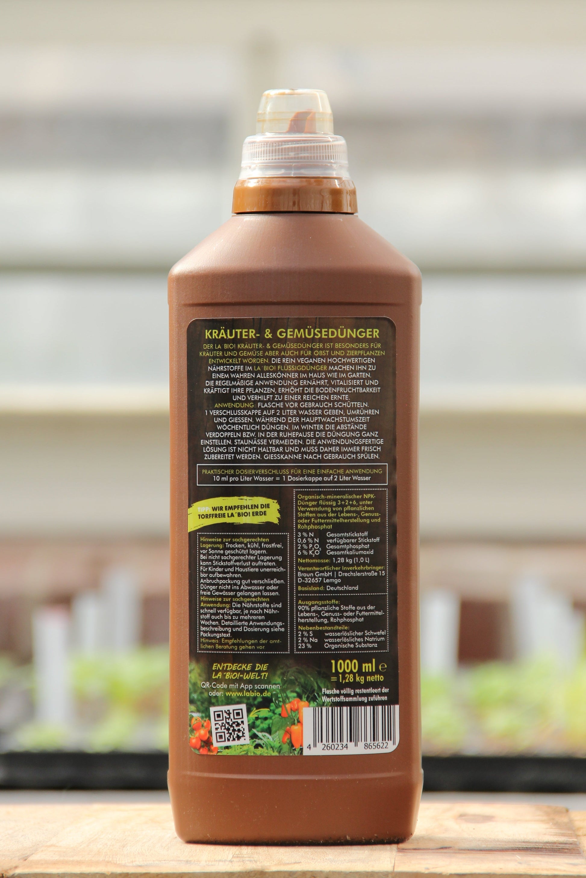 Eine braune Plastikflasche La'Bio | Kräuter & Gemüsedünger 1 l von LaBio mit braunem Verschluss, von hinten gesehen. Das deutsche Etikett zeigt Anweisungen und Dosierungstabellen. Im unscharfen Hintergrund sind grüne Pflanzen zu sehen.