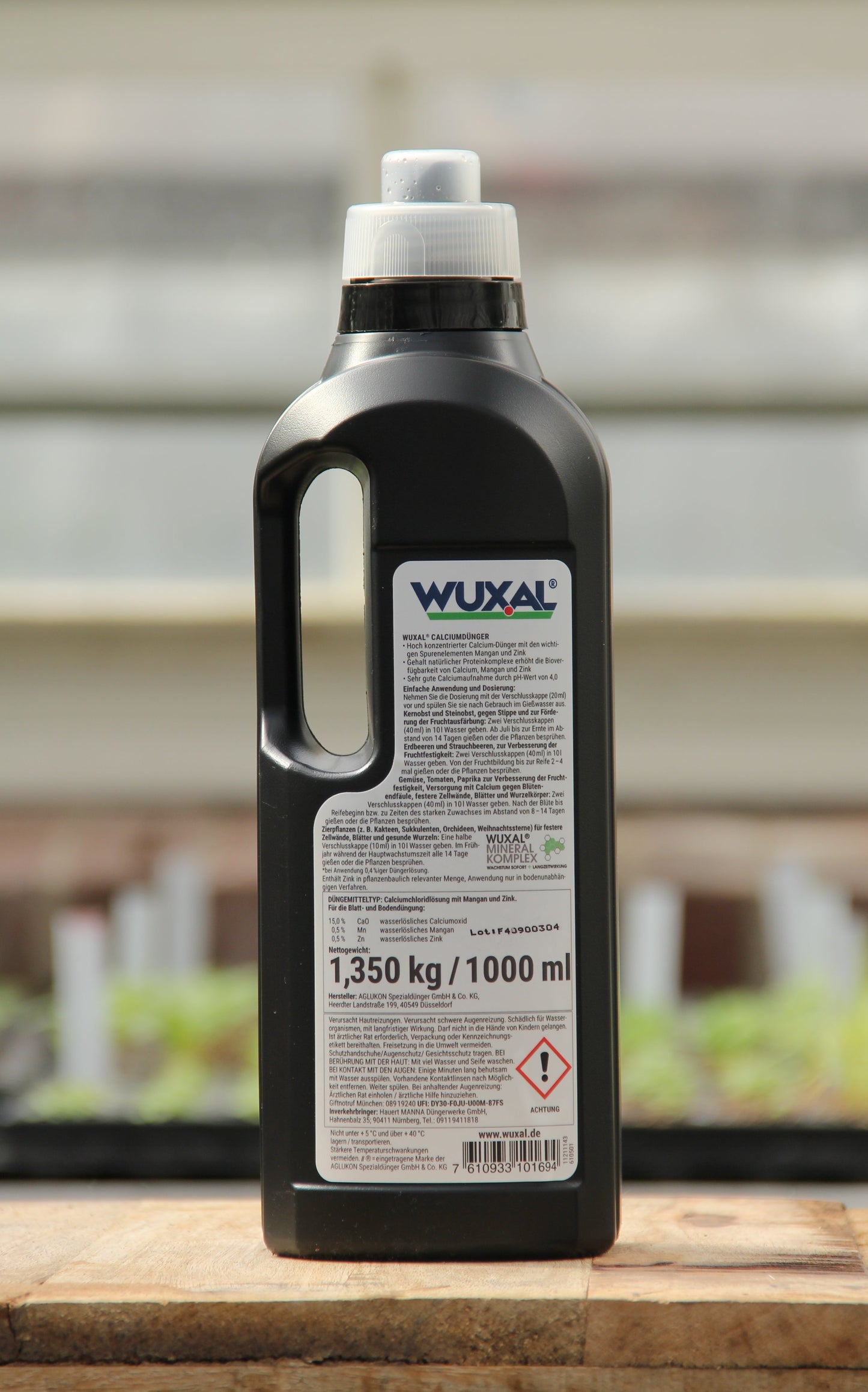 Eine schwarze 1-Liter-Kunststoffflasche von Wuxal-Kalkdünger für Obstbäume, Gemüse und Zierpflanzen mit weißem Deckel, Griff und Etikett mit Produktinformationen und Warnhinweisen steht auf einer Holzfläche mit unscharfem Hintergrund.