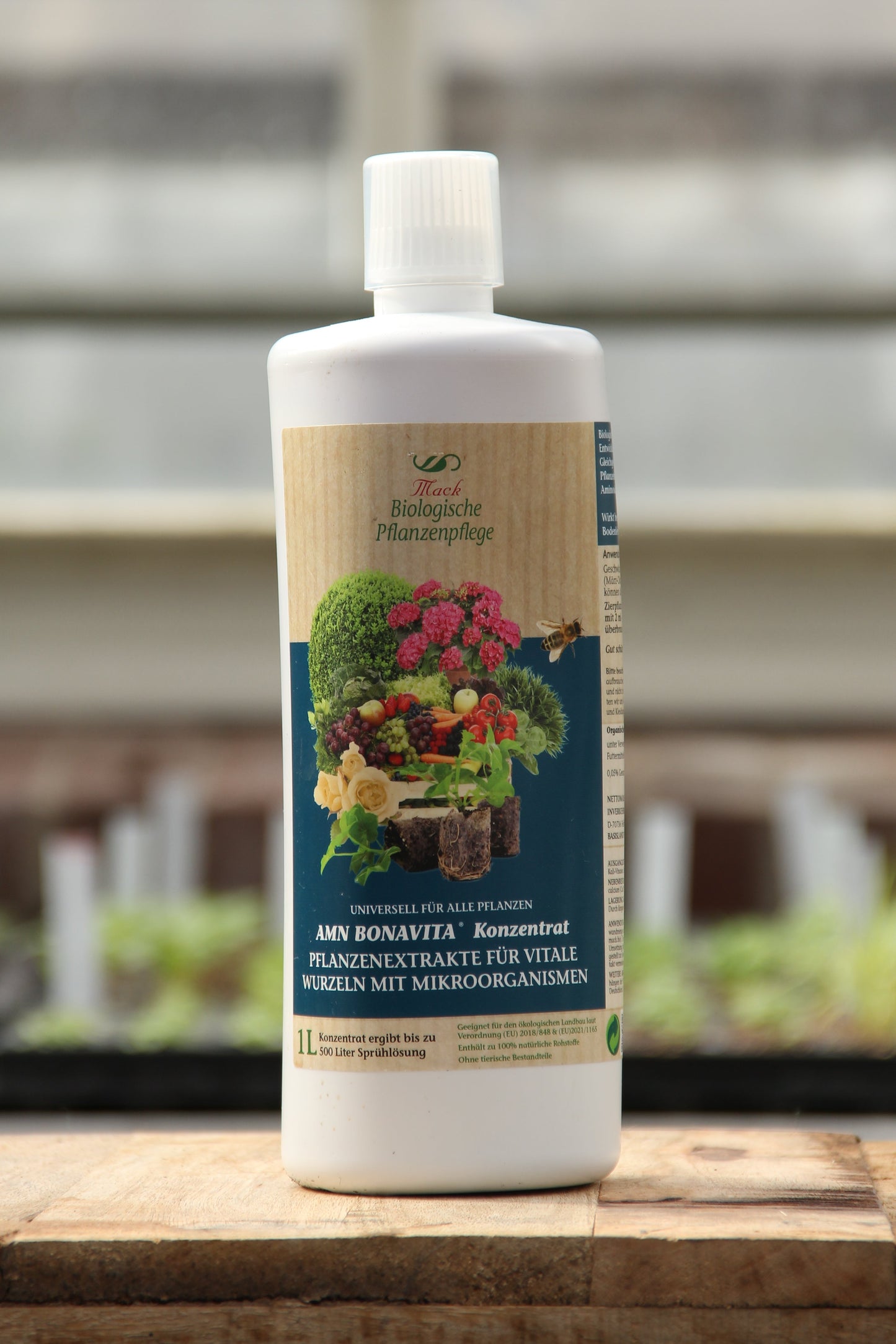 Eine weiße 1-Liter-Flasche AMN 'Bona Vita' Konzentrat mit Mikronährstoffen mit deutscher Aufschrift und Abbildungen von Pflanzen, Früchten und Gemüse steht auf einer hölzernen Fläche mit unscharfem Hintergrund.