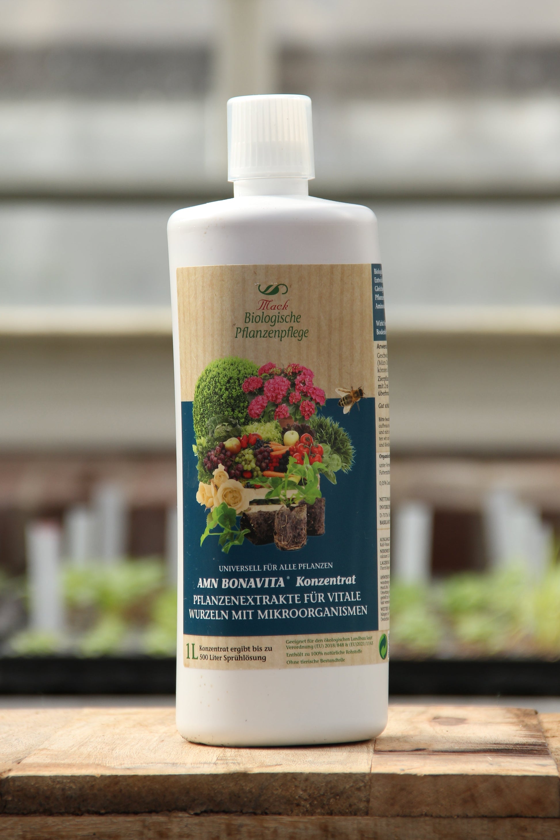 Eine weiße 1-Liter-Flasche AMN 'Bona Vita' Konzentrat mit Mikronährstoffen mit deutscher Aufschrift und Abbildungen von Pflanzen, Früchten und Gemüse steht auf einer hölzernen Fläche mit unscharfem Hintergrund.