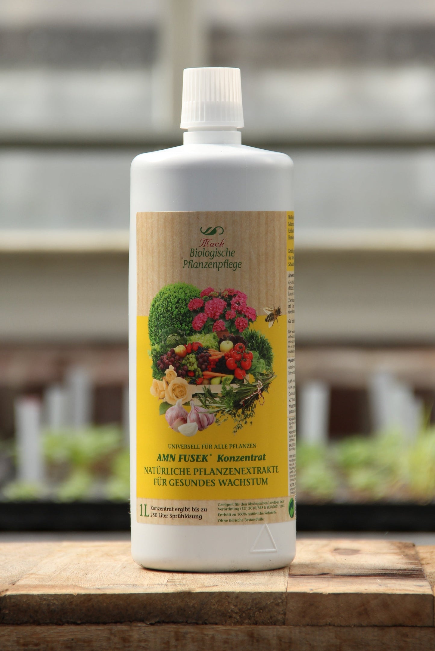 Eine weiße 1L AMN | 'Fusek' Flasche mit biologischem Pflanzenhilfsmittel-Konzentrat mit einem gelben Etikett mit grünen Sträuchern und bunten Blumen, die natürliche Mikroorganismen betonen. Es ruht auf Holz mit einem verschwommenen Hintergrund.