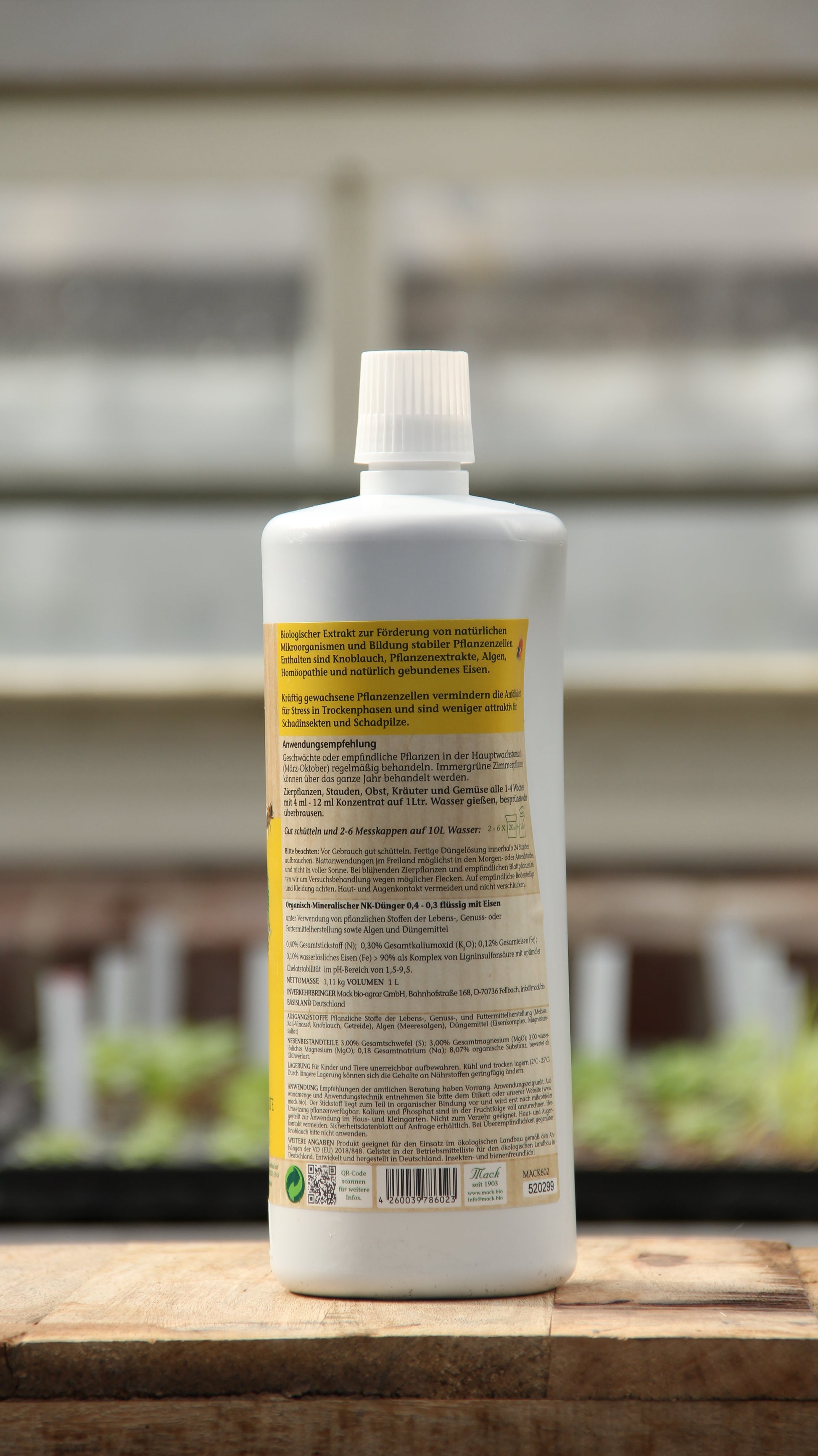 Eine weiße Kunststoffflasche mit weißem Verschluss und gelbem deutschem Etikett mit der Aufschrift "AMN | 'Fusek' für kräftige Pflanzenzellen 1 l" von AMN steht auf einer hölzernen Oberfläche, mit verschwommenen Pflanzen und Gartentabletts im Hintergrund.