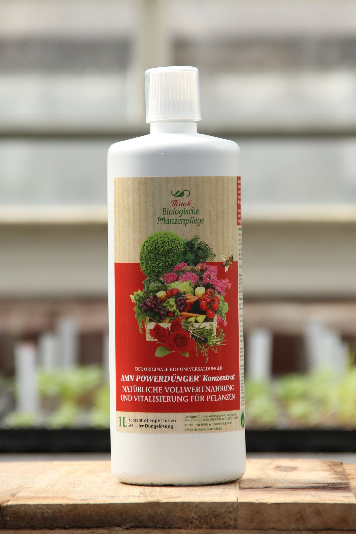 Eine weiße 1-Liter-Flasche AMN | 'Powerdünger' organischer Dünger mit einem roten Etikett, auf dem Gemüse, Früchte und Kräuter abgebildet sind, steht auf einer Holzfläche vor einem unscharfen Hintergrund im Freien.