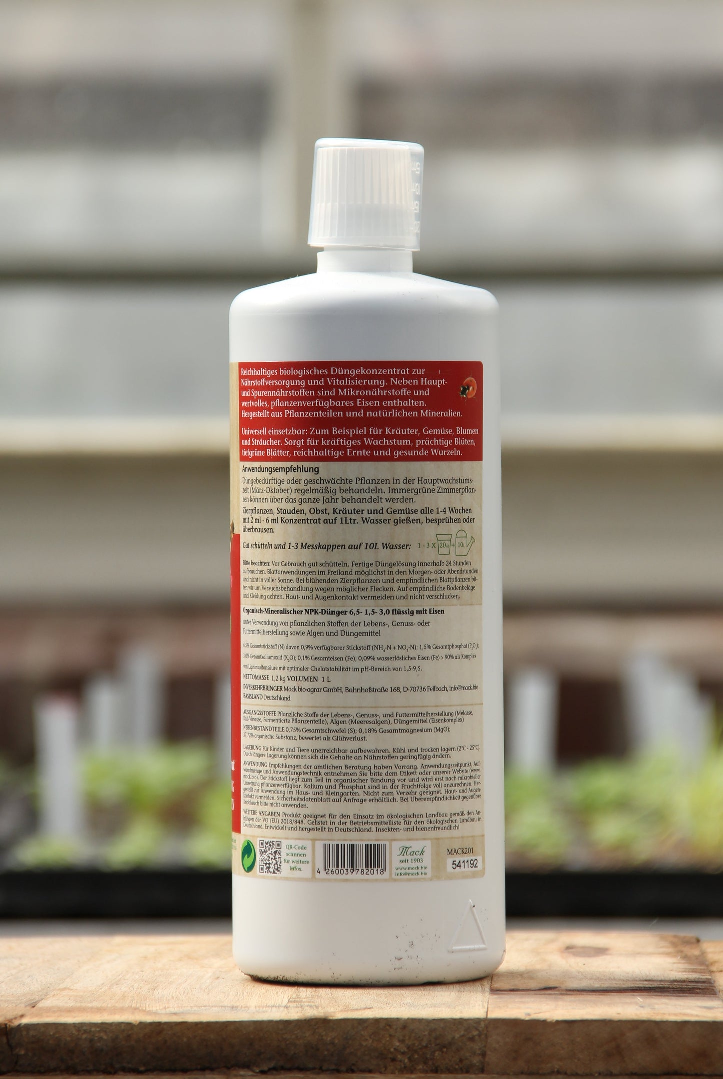 Eine weiße 1-Liter-Plastikflasche von AMN | 'Powerdünger' mit rotem Etikett und weißem Deckel steht auf einer Holzfläche. Auf der Rückseite steht ein ausführlicher deutscher Text über organischen Dünger. Im Hintergrund sind unscharfe Pflanzen und ein Gewächshaus zu sehen.