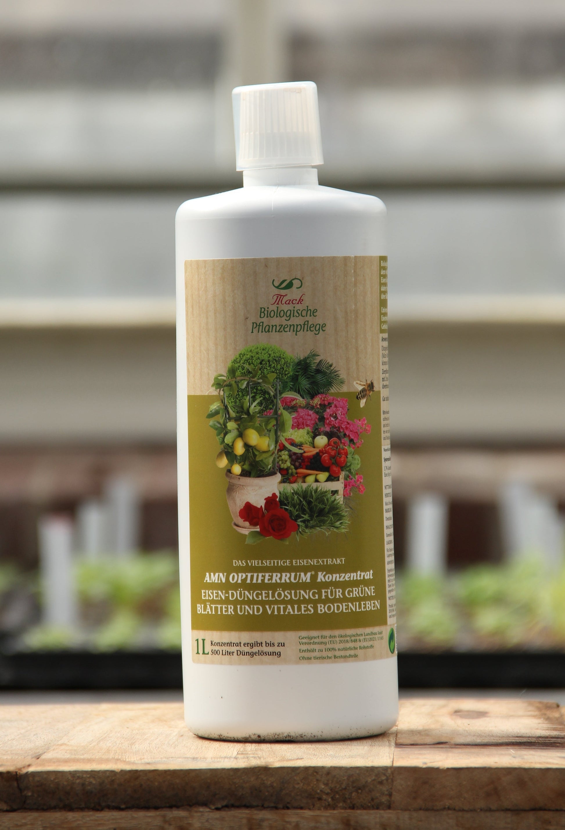 Eine weiße 1-Liter-Flasche AMN 'Optiferrum' organischer Spurennährstoffdünger und Bodenvitalstoff mit grün-gelbem Etikett, auf dem Pflanzen abgebildet sind, auf Holz sitzend, mit verschwommenen Pflanzen und Töpfen im Hintergrund.