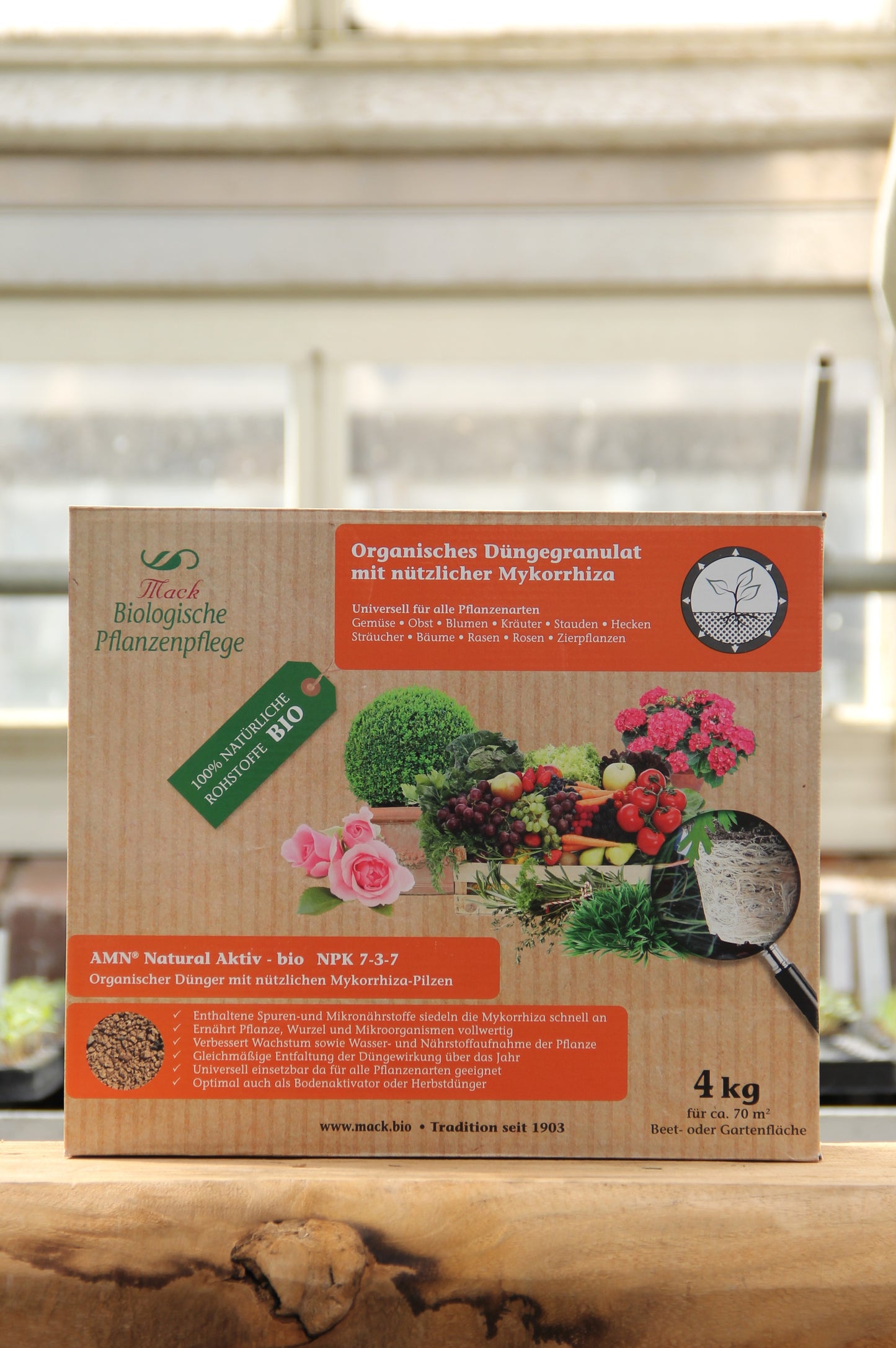 Eine Pappschachtel mit 4 kg organischem Dünger von AMN Natural Aktiv bio" steht auf einer Fensterbank, mit Pflanzen und Gemüse auf der Verpackung mit deutschem Etikett, hinter der das Sonnenlicht hereinscheint.