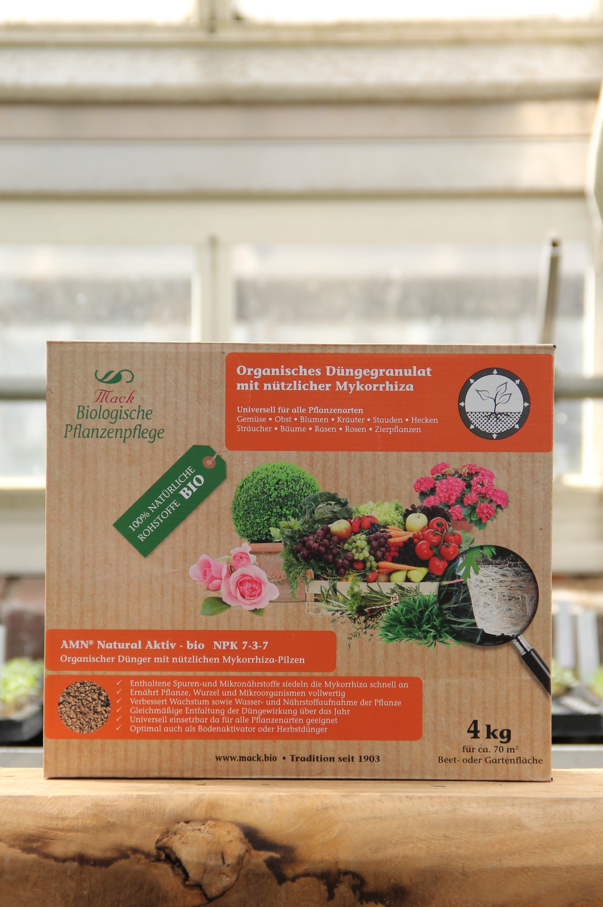 Eine Pappschachtel mit 4 kg organischem Dünger von AMN Natural Aktiv bio" steht auf einer Fensterbank, mit Pflanzen und Gemüse auf der Verpackung mit deutschem Etikett, hinter der das Sonnenlicht hereinscheint.