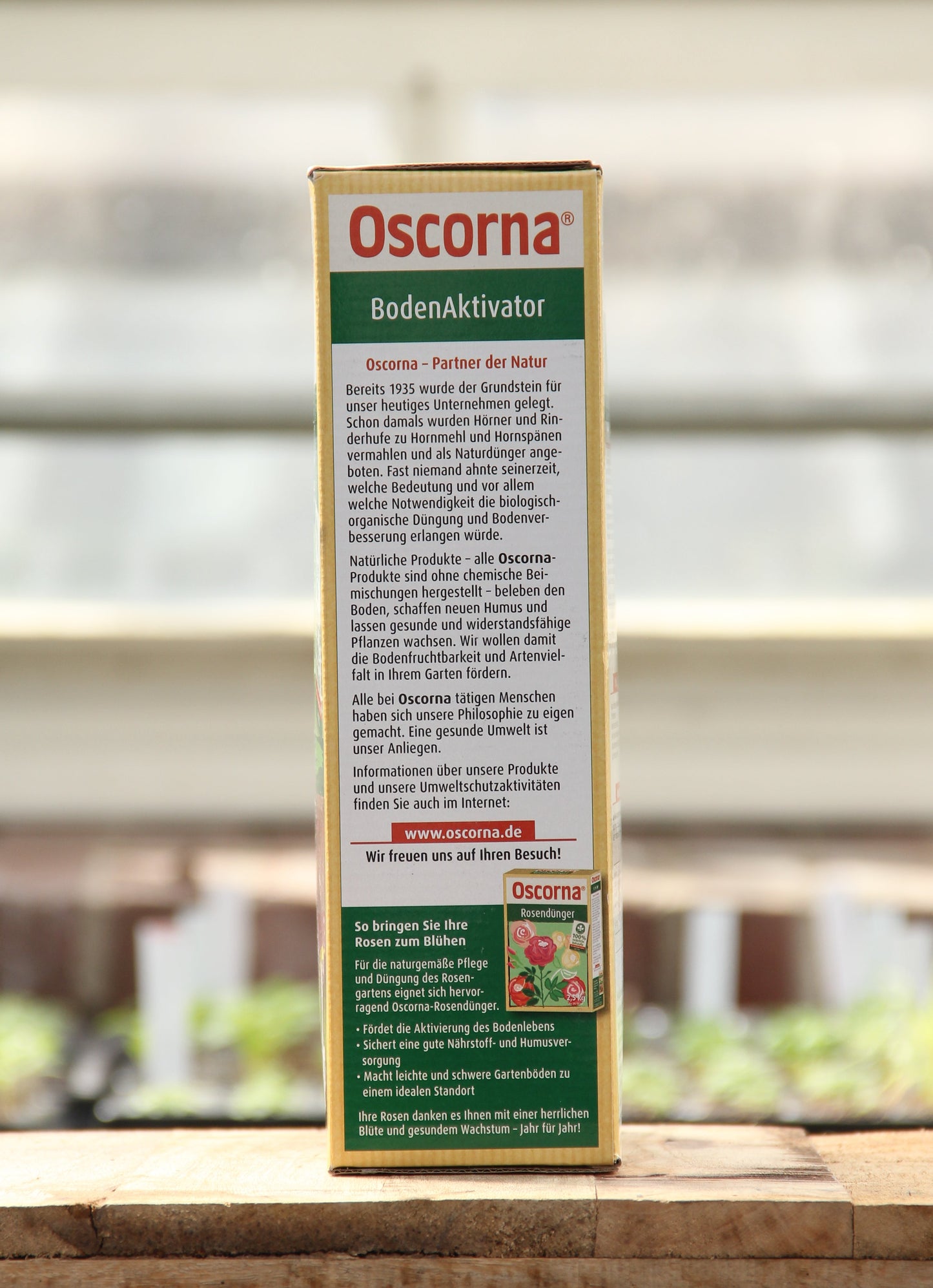 Eine 3 kg Kiste Oscorna | Boden Aktivator steht aufrecht auf Holz und unterstreicht seine Vorteile für das Bodenleben und das Pflanzenwachstum. Auf der Rückseite sind detaillierte Produktinformationen in deutscher Sprache vor einem sanft verschwommenen Hintergrund abgebildet.