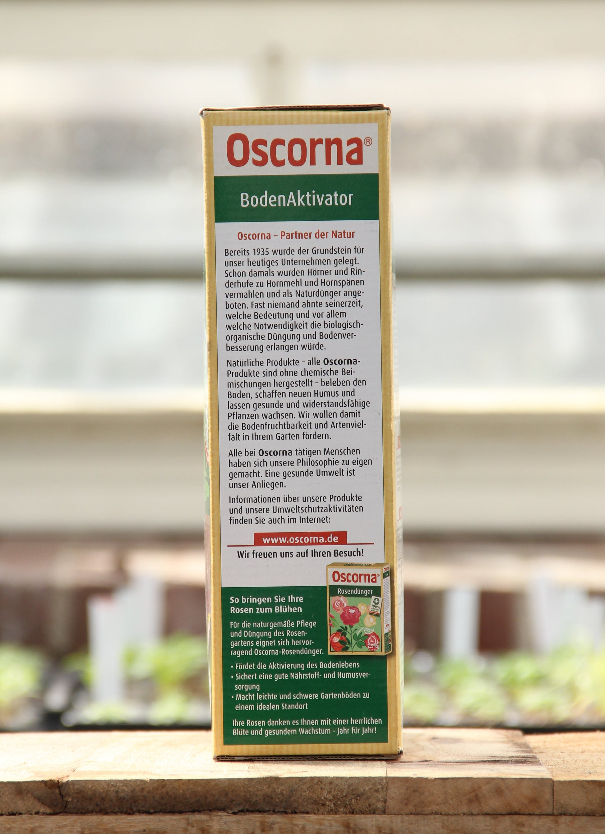 Eine 3 kg Kiste Oscorna | Boden Aktivator steht aufrecht auf Holz und unterstreicht seine Vorteile für das Bodenleben und das Pflanzenwachstum. Auf der Rückseite sind detaillierte Produktinformationen in deutscher Sprache vor einem sanft verschwommenen Hintergrund abgebildet.