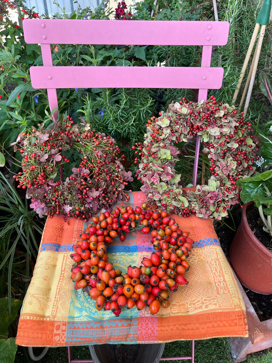 Ein rosafarbener Metallstuhl mit bunter Tischdecke zeigt drei handgefertigte Kränze - eine ideale Inspiration für den "Di. 29.09.2026 | 11.00-12.30 WORKSHOP Herbstliches Türkranzbinden" der Königlichen Gartenakademie inmitten von üppigem Gartengrün.