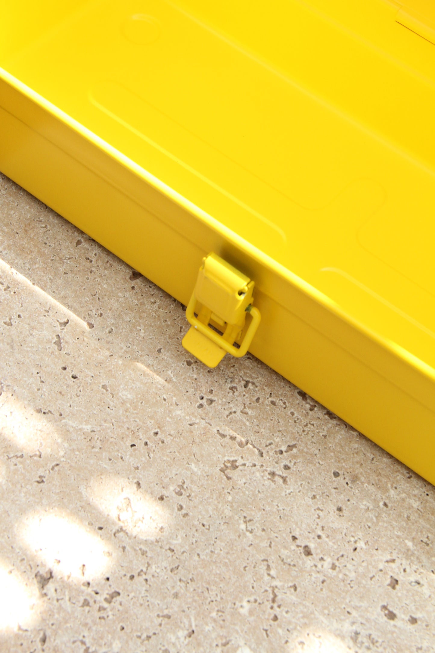 Nahaufnahme eines leuchtend gelben Metallverschlusses an der Werkzeugkiste Niwaki | 'Y-Type Toolbox' (35 x 15 x 10,5 cm), die auf einem strukturierten, hellen Stein mit gesprenkeltem Sonnenlicht ruht.