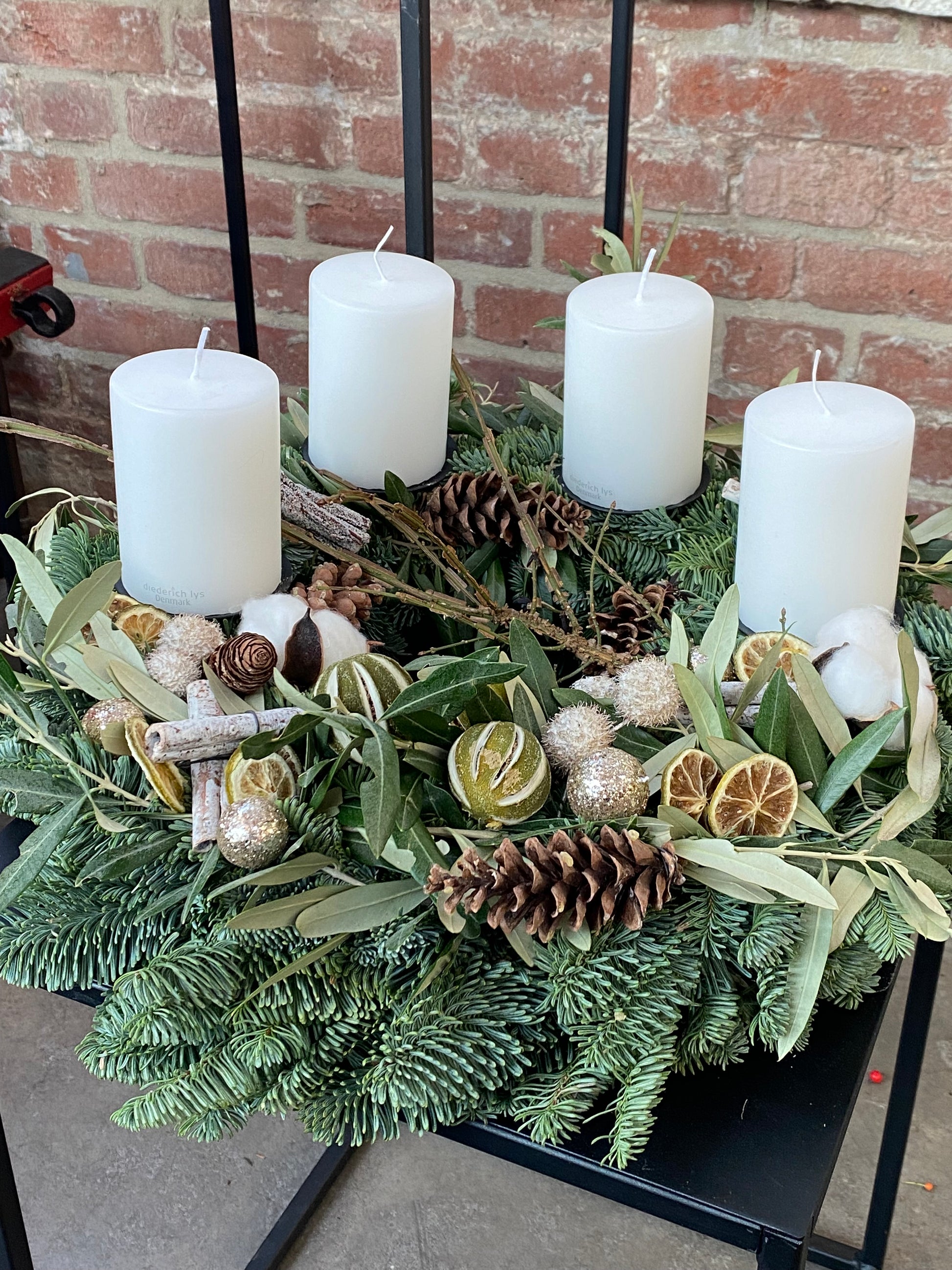 Lassen Sie sich inspirieren für Ihr eigenes Do. 26.11.2026 | 10:00-12:00 WORKSHOP Adventskranzbinden by Königliche Gartenakademie-ein festlicher Kranz mit weißen Kerzen, Tannenzapfen, getrockneten Zitrusfrüchten, Zimt, Baumwolle & Glitter auf Immergrün vor einer Backsteinwand!.