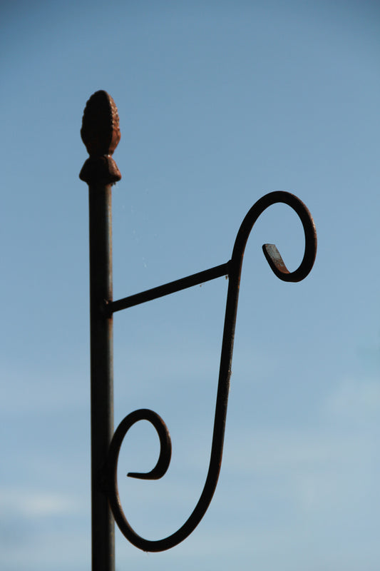 Ein Hirsch-Laternenstab „Bino Pinie“, erhältlich in den Längen 1,7 oder 2,0 Meter, gefertigt aus schwarzem Metall mit dekorativen Schnörkeln, ragt vor einem strahlend blauen Himmel empor und erinnert an die elegante Form eines Rankstabs.