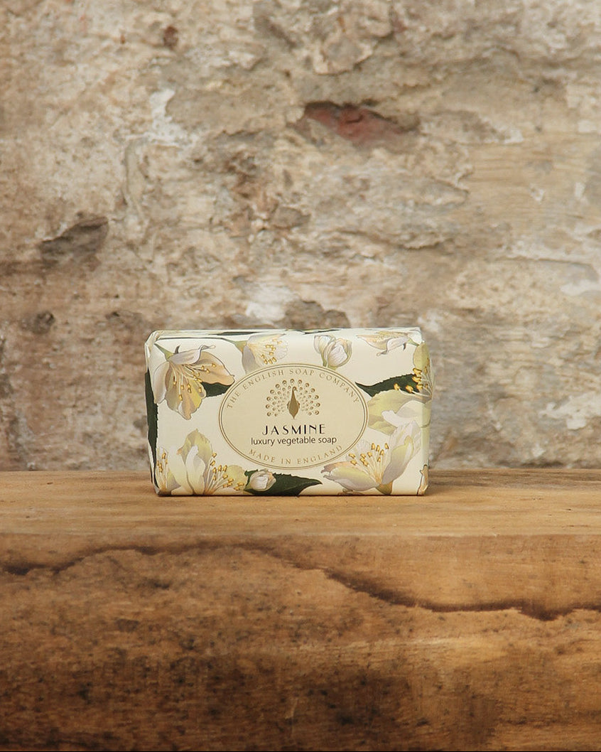 Ein Riegel ESC | 'Jasmin' Pflanzliche Handseife aus seidigen Blütenakkorden von The English Soap Company ruht auf rustikalem Holz, seine plastikfreie Verpackung ergänzt die verwitterte Steinwand dahinter.