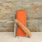 Das Niwaki | 'Kotoh X Niwaki Knife' Taschenmesser mit Holzgriff und teils geöffneter Edelstahlklinge lehnt an einer orangefarbenen Lederschutzhülle auf Stein-ideal für Gartenarbeit-vor einer strukturierten Ziegelwand.