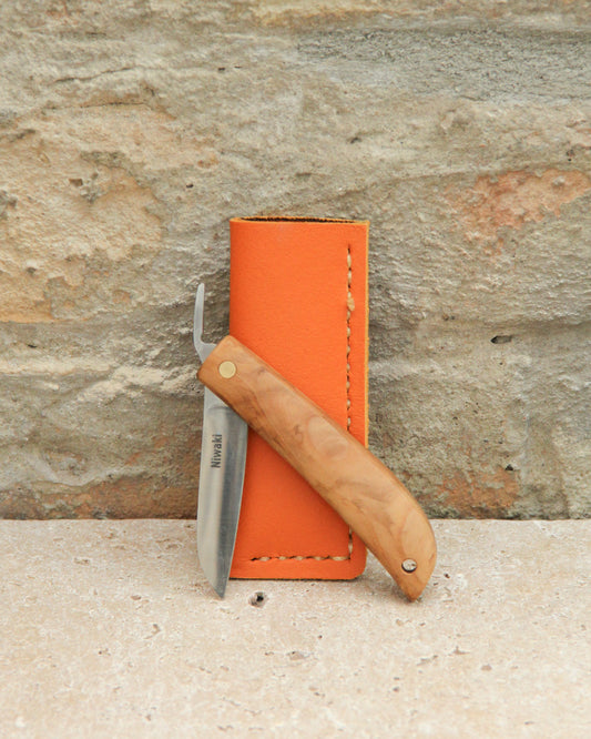 Das Niwaki | 'Kotoh X Niwaki Knife' Taschenmesser mit Holzgriff und teils geöffneter Edelstahlklinge lehnt an einer orangefarbenen Lederschutzhülle auf Stein-ideal für Gartenarbeit-vor einer strukturierten Ziegelwand.