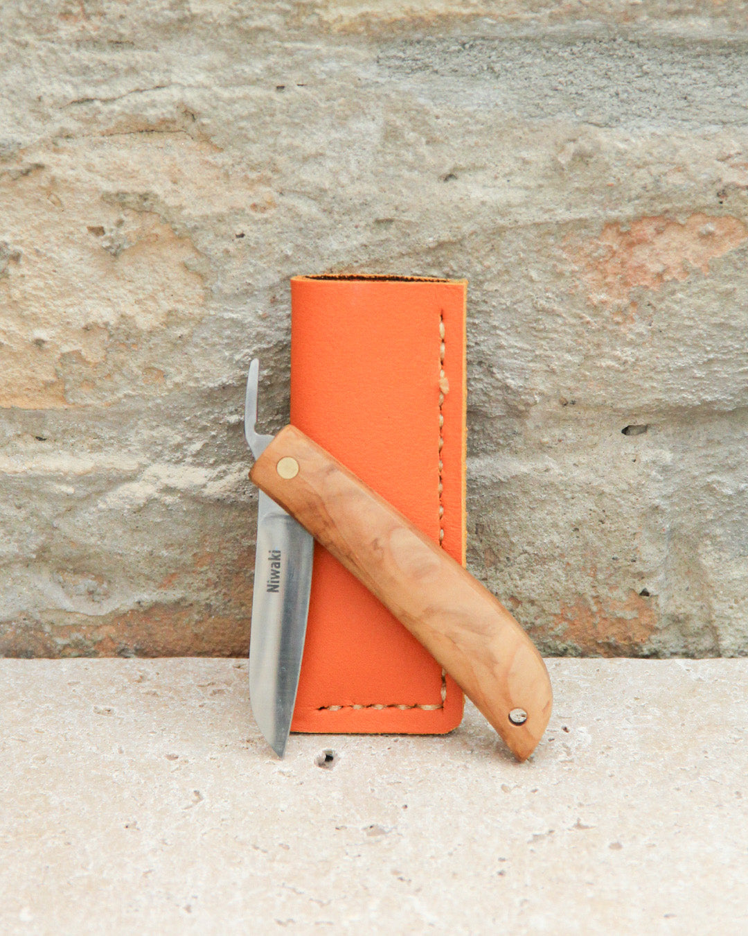 Ein Niwaki | Das „Kotoh X Niwaki Knife“ – ein Taschenmesser mit Lederschutzhülle – lehnt mit seinem Holzgriff und der gebogenen Klinge an einer orangefarbenen Lederschutzhülle vor einer strukturierten Steinwand – ideal für präzise Bonsai-Schnittarbeiten und Gartenarbeiten.