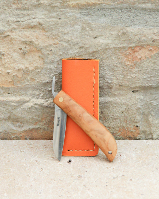 Ein Niwaki | Das „Kotoh X Niwaki Knife“ – ein Taschenmesser mit Lederschutzhülle – lehnt mit seinem Holzgriff und der gebogenen Klinge an einer orangefarbenen Lederschutzhülle vor einer strukturierten Steinwand – ideal für präzise Bonsai-Schnittarbeiten und Gartenarbeiten.