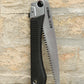 Die Niwaki | „GR 210 Folding Saw“ Klappsäge mit schwarzem, strukturiertem Griff und teilweise freiliegendem silbernem Sägeblatt steht aufrecht vor einer beigen Backsteinmauer – ein ideales Werkzeug von Niwaki für präzise Schnitte in jeder Außenumgebung.