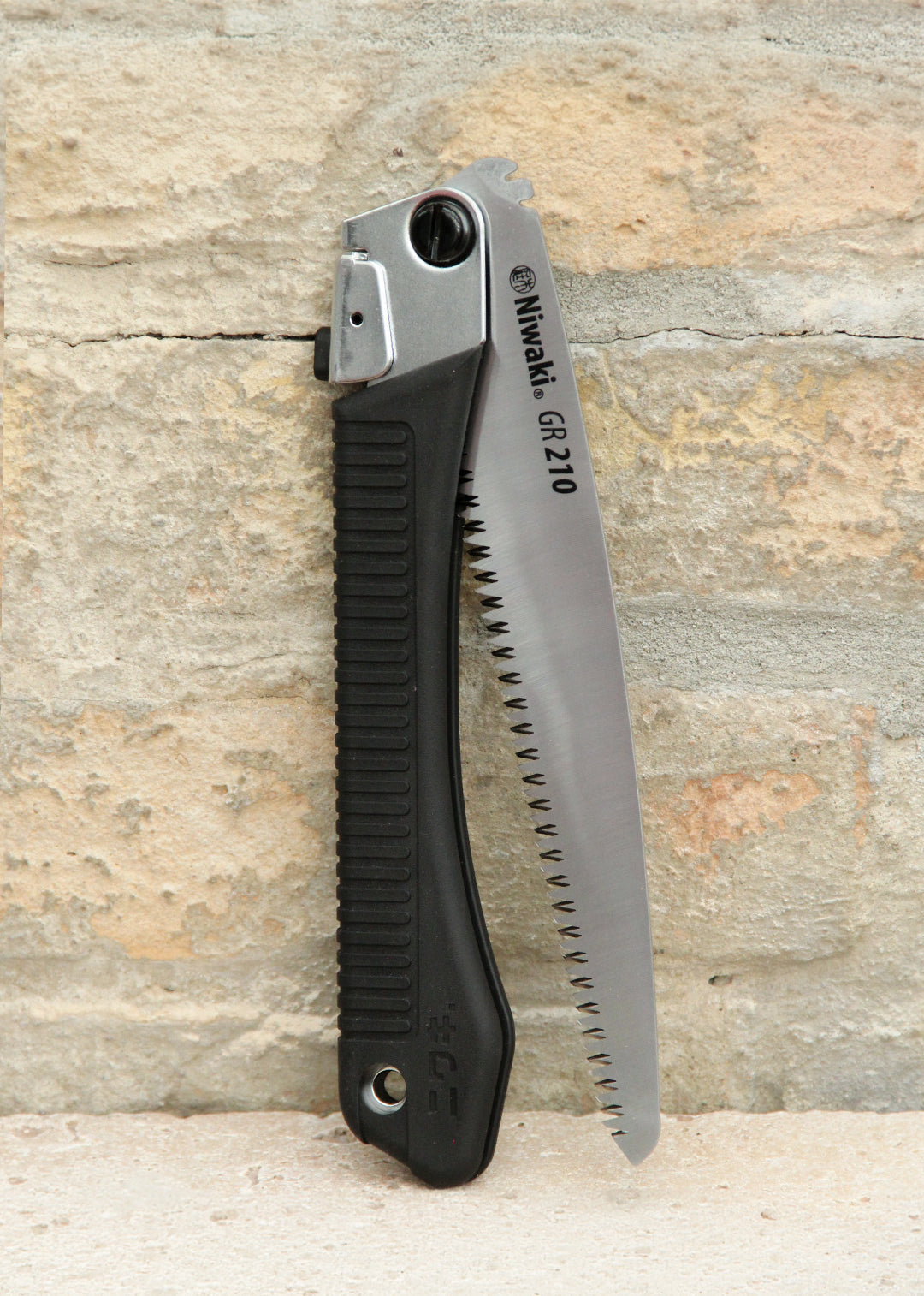 Die Niwaki | „GR 210 Folding Saw“ Klappsäge mit schwarzem, strukturiertem Griff und teilweise freiliegendem silbernem Sägeblatt steht aufrecht vor einer beigen Backsteinmauer – ein ideales Werkzeug von Niwaki für präzise Schnitte in jeder Außenumgebung.