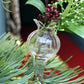 Ein Des Pots Filigrane Mini Klemmvasen aus dem 6er Set, gefüllt mit grünen Blättern und roten Beeren, hängt zwischen Tannennadeln vor einem verschwommenen roten Hintergrund.