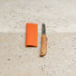 Das Niwaki | „Kotoh X Niwaki Knife“ Taschenmesser mit Lederschutzhülle – ein Klappmesser mit Holzgriff, perfekt für Gartenarbeiten und präzise Bonsai-Schnitte – liegt aufgeklappt neben seiner orangefarbenen Lederschutzhülle auf einer hellbeigen Steinfläche. Dieses Modell von Niwaki vereint Funktionalität und elegantes Design und ist damit die ideale Wahl für Gartenliebhaber.