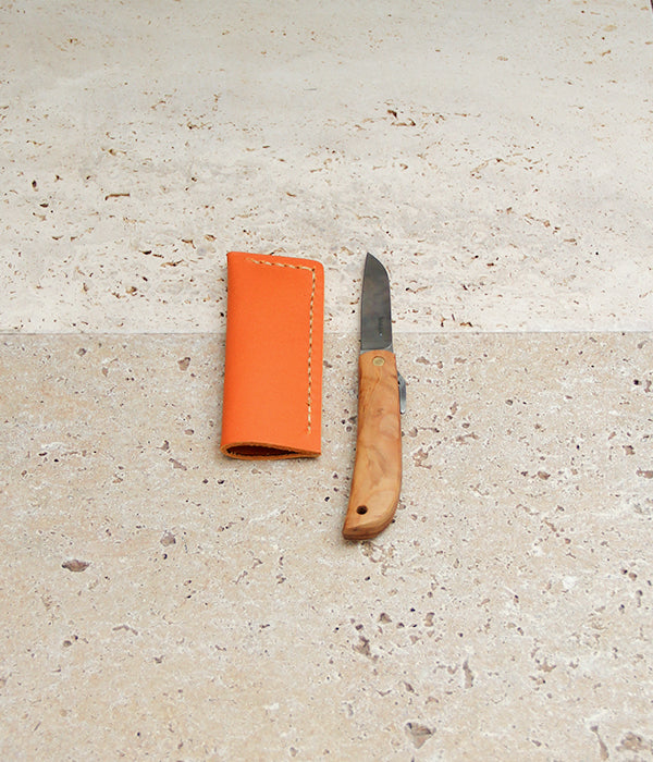 Das Niwaki | „Kotoh X Niwaki Knife“ Taschenmesser mit Lederschutzhülle – ein Klappmesser mit Holzgriff, perfekt für Gartenarbeiten und präzise Bonsai-Schnitte – liegt aufgeklappt neben seiner orangefarbenen Lederschutzhülle auf einer hellbeigen Steinfläche. Dieses Modell von Niwaki vereint Funktionalität und elegantes Design und ist damit die ideale Wahl für Gartenliebhaber.