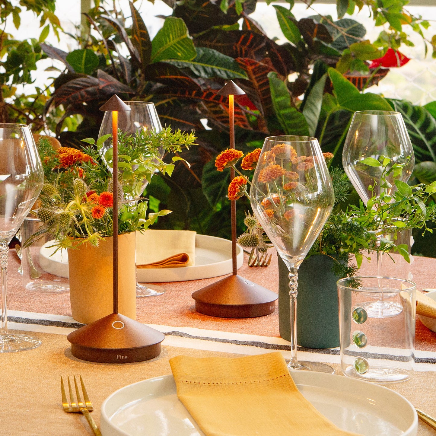 Ein gedeckter Esstisch mit leeren Tellern, Weingläsern und einem Wasserglas ist mit kleinen Vasen mit orangefarbenen Blumen, Grünzeug und zwei modernen Tischleuchten von Zafferano | Pina 'Corten' dekoriert. Üppige grüne Pflanzen füllen den Hintergrund.