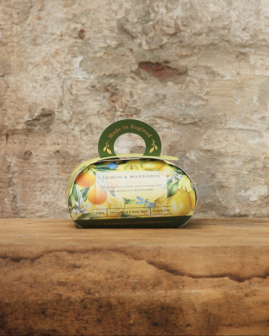Eine dekorative ovale Geschenkschachtel mit der nach Zitrusfrüchten duftenden pflanzlichen Handseife "Lemon & Mandarin" von The English Soap Company ESC, verziert mit Zitronen- und Mandarinenbildern und einem kleinen Griff, steht auf einer hölzernen Oberfläche vor einer rustikalen Steinwand.