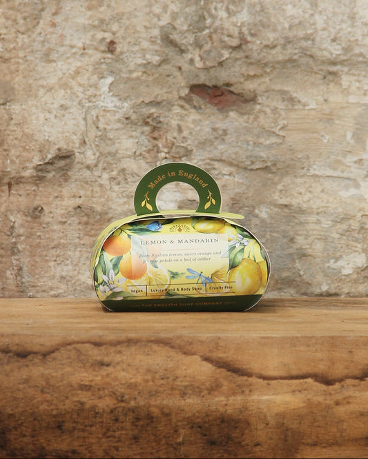 Eine dekorative ovale Geschenkschachtel mit der nach Zitrusfrüchten duftenden pflanzlichen Handseife "Lemon & Mandarin" von The English Soap Company ESC, verziert mit Zitronen- und Mandarinenbildern und einem kleinen Griff, steht auf einer hölzernen Oberfläche vor einer rustikalen Steinwand.