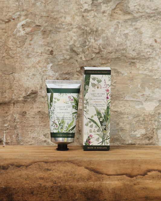 Eine Tube The English Soap Company ESC | 'Lily of the Valley' Handcreme mit Maiglöckchenduft 75 ml ruht neben der dazugehörigen Schachtel auf einer rustikalen Holzfläche mit einer strukturierten Steinwand im Hintergrund.