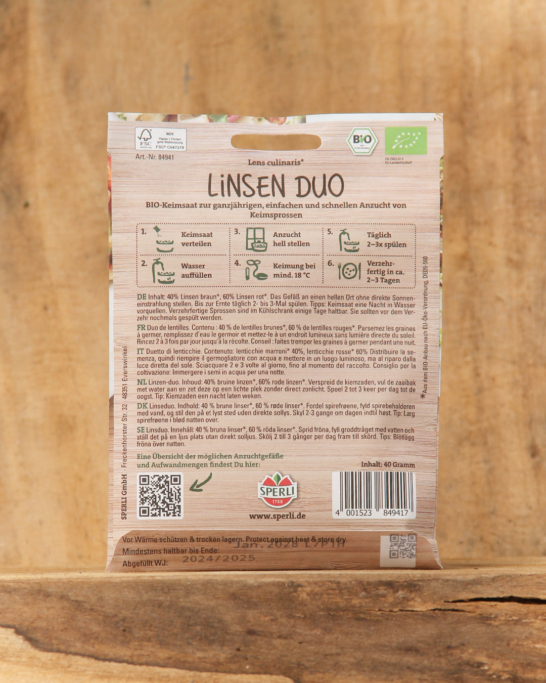 Die Saatgutpackung von Sperli's Bio Keimsprossen Saatgut 'Linsen Duo' steht aufrecht auf einem Holzständer. Auf der Rückseite befinden sich deutsche Pflanzanweisungen, Beschreibungen, Symbole und ein Strichcode; der Markenname Sperli's ist deutlich zu erkennen.