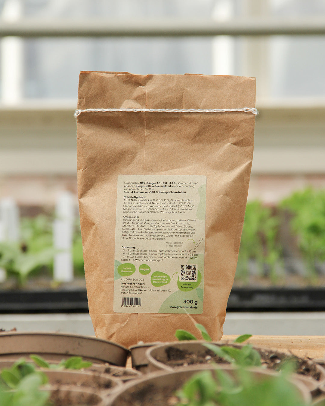 A Green Mundo 'Luzi' 300g vegane Düngestäbchen-Tüte für Zimmer- und Topfpflanzen steht auf Holz. Im Hintergrund unscharfe grüne Pflanzen, das Label zeigt Text, Icons und einen QR-Code.
