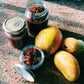Drei Gläser „Wilkin & Sons | Tiptree Chutney Mango“ (220 g) stehen auf einer Granitarbeitsplatte neben drei ganzen, reifen Mangos. Zwei Gläser des Wilkin & Sons-Chutneys sind verschlossen, während eines offen steht und den Blick auf das stückige Mango-Chutney im Inneren freigibt. Sonnenlicht strömt herein und wirft sanfte Schatten auf die Anordnung.