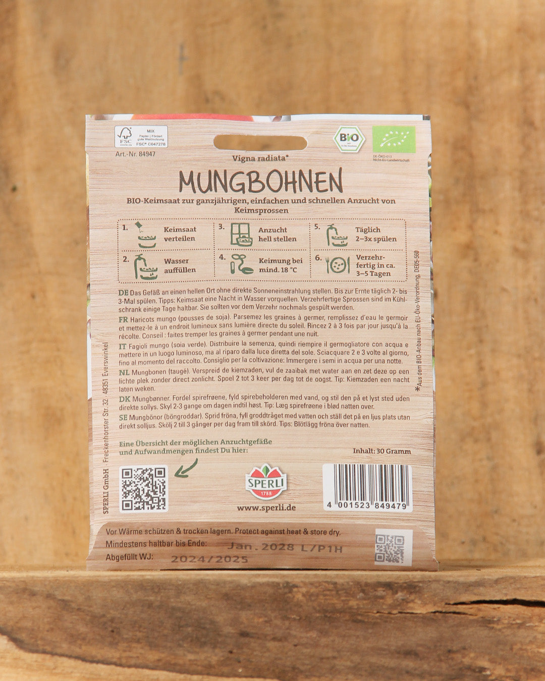 Die Rückseite der Bio Keimsprossen Saatgut 'Mungbohnen' von Sperli zeigt eine deutsche Aussaatanleitung, Icons, einen QR-Code, das Logo von Sperli, einen Barcode und Nachhaltigkeitsetiketten - alles auf einem Holzuntergrund.