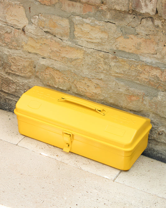 Eine gelbe Niwaki 'Y-Type Toolbox' Werkzeugbox (35 x 15 x 10,5 cm) mit Griff und Verschluss liegt auf einer hellen Steinfläche vor einer rustikalen Ziegelwand.