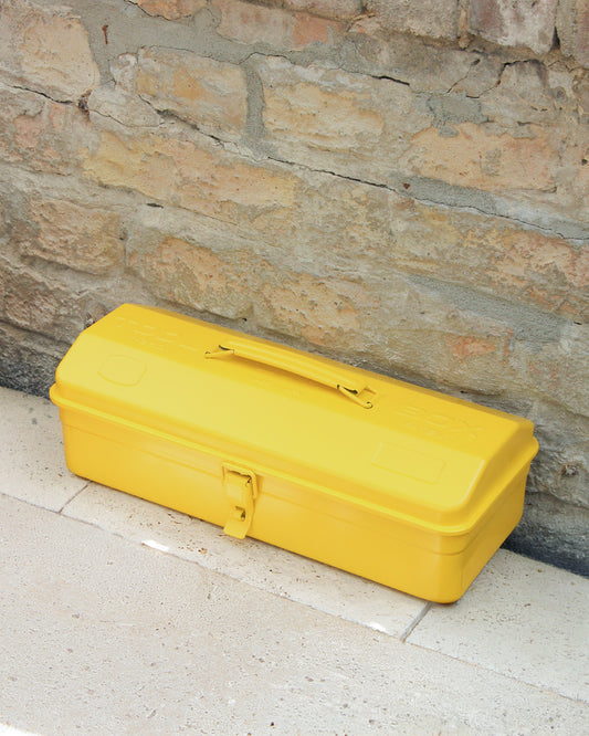Eine geschlossene, gelbmetallfarbene Niwaki | „Y-Type Toolbox“ Werkzeugbox (35 x 15 x 10,5 cm) mit Griff steht auf einer hellen Steinfläche vor einer rustikalen Backsteinmauer und ist bereit, all Ihre wichtigen Utensilien für die Gartenarbeit aufzunehmen.