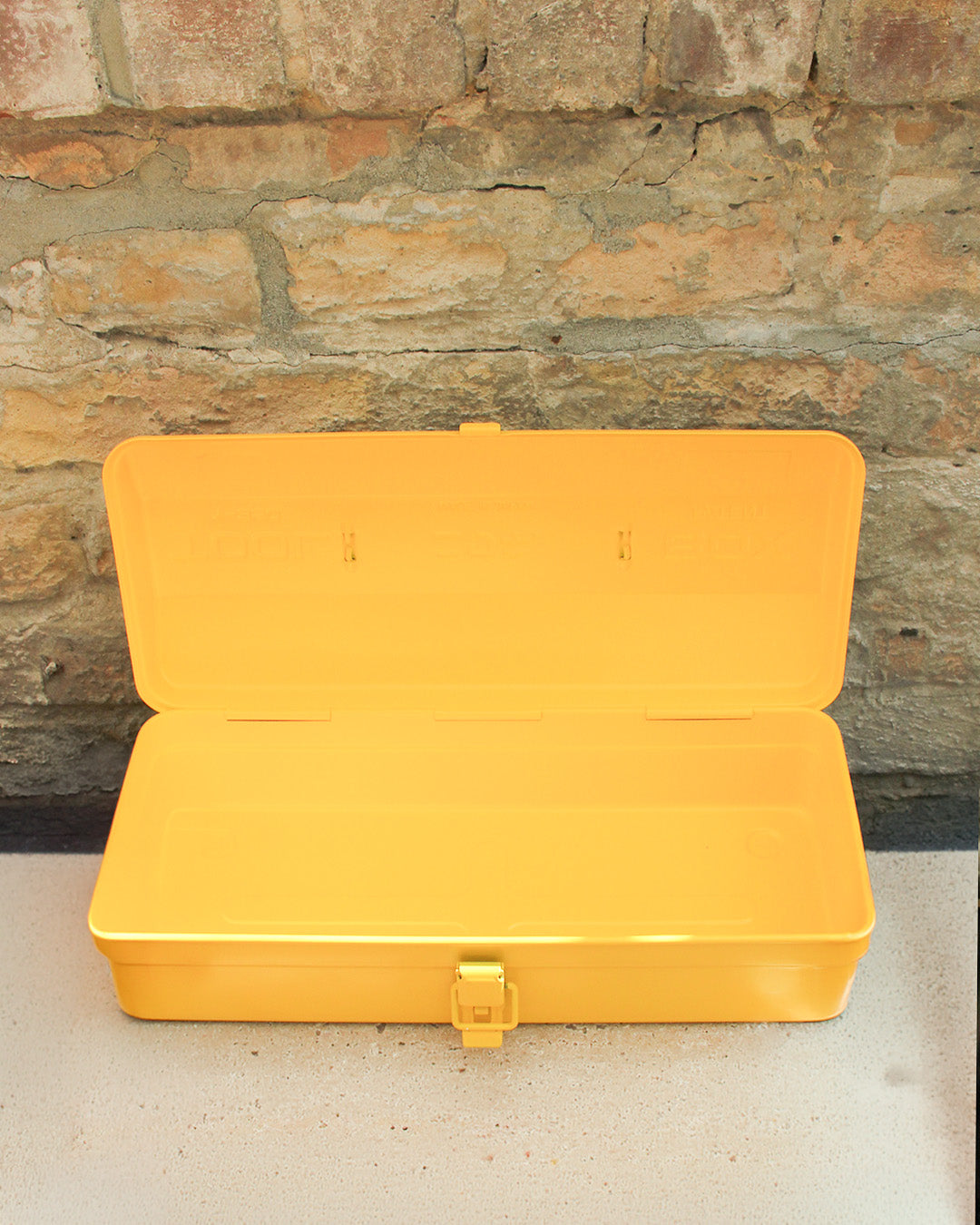 Die Niwaki | 'Y-Type Toolbox' Werkzeugbox (35 x 15 x 10,5 cm) von Niwaki steht mit offenem Deckel und leerem Innenraum auf einer hellen Fläche vor einer rustikalen Backsteinwand; vorn ist der Verschluss zu sehen.