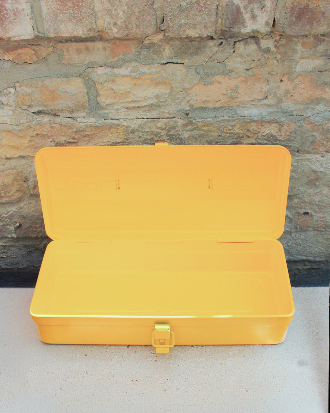Eine leuchtend gelbe Niwaki | „Y-Type Toolbox“ Werkzeugbox (35 x 15 x 10,5 cm) steht offen auf einer weißen Fläche vor einer strukturierten Ziegelwand. Diese robuste Metallbox von Niwaki, die sich ideal zur Aufbewahrung von Gartengeräten eignet, ist leer und verfügt an der Vorderseite über einen schlichten Verschluss.