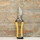 Ein Paar Niwaki | 'Higurashi GR Secateurs' Gartenschere von Niwaki mit gelben und schwarzen Griffen steht aufrecht vor einer strukturierten hellbraunen Steinwand.