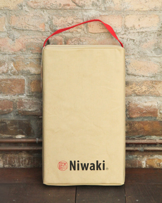 Eine rechteckige beigefarbene Tasche mit rotem Henkel und dem Niwaki-Logo in Schwarz und Rot steht aufrecht an einer Backsteinmauer - ideales Gärtner-Zubehör für Ihre Gartenarbeit oder als Ergänzung zu Ihrem Niwaki | Kniekissen von Niwaki.