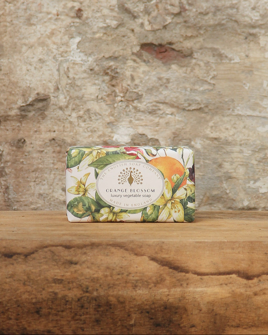 Eine dekorative Tafel von ESC | 'Orange Blossom' pflanzliche Handseife mit fruchtigem Orangenblütenduft von The English Soap Company, verziert mit Blumen- und Fruchtmotiven, liegt auf rustikalem Holz vor einer Steinwand.